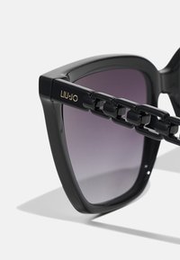 Occhiali da sole neri con montatura spessa, lenti viola sfumate e un design a catena sul tempio. Presentano il nome del marchio "LIU-JO."