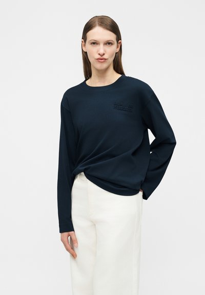 Γυναίκα με καστανά ίσια μαλλιά φοράει σκούρο μπλε φούτερ και λευκό παντελόνι, στέκεται μπροστά σε μια απλή ανοιχτογκρι φόντο.