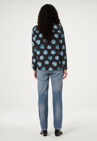 Schwarze Bluse mit türkisfarbenem Blumenmuster, langen Ärmeln und rundem Ausschnitt, kombiniert mit lässigen, hellblauen Jeans und schwarzen High Heels.