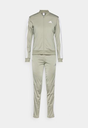 Olive spalvos treniruočių kostiumas su užtrauktuku, atitinkantys kelnių, su baltais šoninėmis juostelėmis ir „Adidas“ logotipu kairėje krūtinės ir šlaunies pusėje.