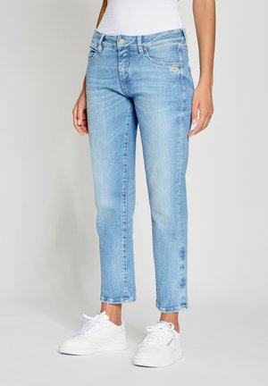 Frau trägt hellblaue Jeans mit geradem Bein und weiße Sneakers, steht vor einem einfarbigen grauen Hintergrund.
