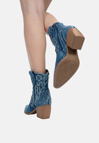 Botas de tobillo de ante azul con diseños bordados de flores y estrellas, cierre con cremallera y tacones cuadrados marrones. Suela de goma texturizada.