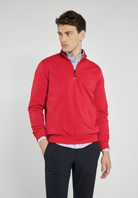 Czerwony sweter z kieszenią i ćwierć zamkiem, z wykończonym ściągaczem kołnierza i mankietów, wykonany z gładkiego, lekkiego materiału. Widać czarny detal z logo.