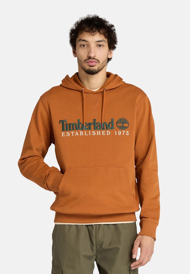 Hoodie marron avec une poche kangourou, présentant un graphisme vert et blanc "Timberland Established 1973" sur le devant. Tissu en mélange de coton.