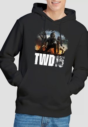 Person trägt schwarzen Kapuzenpullover mit Grafik von drei Charakteren in Aktionsposen und Schriftzug "TWD 15" auf der Vorderseite.