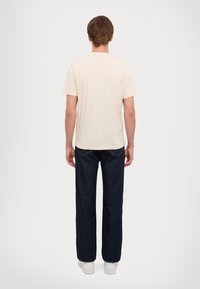 T-shirt di cotone beige chiaro con collo rotondo, abbinata a jeans di denim blu scuro. Le scarpe sono sneakers bianche, viste da dietro.
