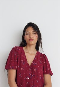 Burgunderfärgad blommig klänning med puffärmar, V-ringning och knappdetaljer. Tyget är lätt och har mönster av röda, blå och gula blommor.