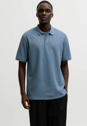 Uomo che indossa una polo a maniche corte azzurro chiaro e pantaloni neri a righe, in piedi con una mano in tasca contro uno sfondo semplice.