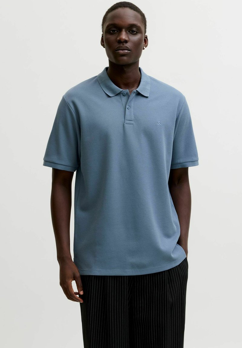 Férfi világoskék rövid ujjú pólóingben és fekete csíkos nadrágban, egyik kezét a zsebébe téve, egyszerű háttér előtt áll.