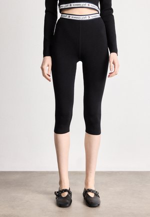 Legging noirs cropped avec une taille haute portant un logo blanc "tommy girl". Texture lisse avec un design ajusté, associés à des chaussures noires.