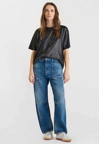 Zwarte, korte mouwen, glanzende top gecombineerd met losse blauwe spijkerbroek met buikzakken; gedragen met witte sneakers op een effen achtergrond.