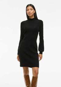 Vestito a maglia nero con collo alto, maniche lunghe e silhouette aderente. Presenta una texture a coste ed è di lunghezza al ginocchio. Indossato con stivali alti marroni.