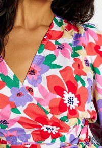 Haut enroulé floral en coton avec des motifs vibrants rouges, roses, violets et verts, doté d'un décolleté en V profond et de manches froncées.
