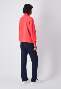 Koraal sweatshirt met hoge kraag, geribbelde manchetten en zoom; gecombineerd met marineblauwe broek; model draagt witte sneakers, gezien vanuit de achterkant.
