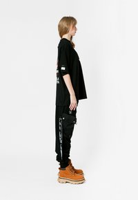 Camiseta negra de gran tamaño con estampado gráfico, combinada con pantalones cargo negros con bolsillos laterales y botas marrones con suelas gruesas.