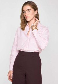 Chemise à rayures rose clair à manches longues, assortie à un pantalon taille haute bordeaux foncé. Des bracelets en or ornent le poignet.