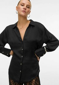 Crna košulja na dugim rukavima s satenskom teksturom, s kragnom i srebrnim detaljima na dugmima. Elegantna s blagim zakrivljenim rubom.
