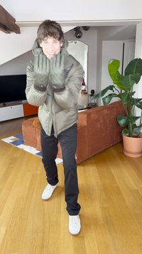 Φουσκωτό μπουφάν πράσινου ελιάς, σκούρο παντελόνι, λευκά αθλητικά παπούτσια και γκρι γάντια. Εσωτερικός χώρος με ξύλινο πάτωμα και ένα μεγάλο φυτό στο παρασκήνιο.