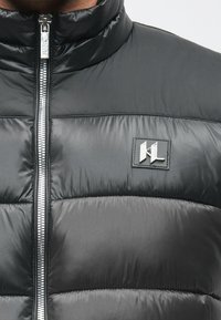 Čierna prešívaná bunda s vysokým golierom, vybavená strieborným zipsom a textúrovaným prešívaným dizajnom. Na prednej strane je výrazná logo nášivka.