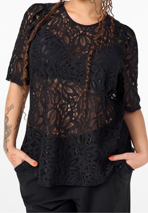 Haut noir en dentelle à manches courtes avec des motifs floraux porté sur la peau, associé à un pantalon noir, mains dans les poches et tatouages visibles sur le bras gauche.