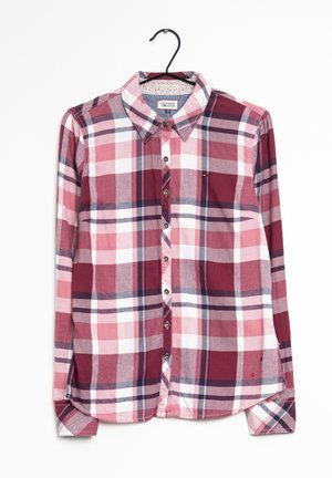 Chemise à manches longues boutonnée avec un motif à carreaux rouge, rose, blanc et noir, suspendue sur un cintre devant un fond blanc.