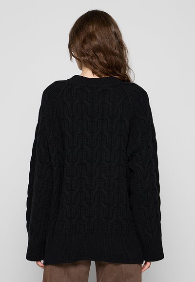 Femme aux cheveux bruns portant un long pull en maille torsadée noir et un pantalon marron, vue de dos sur un fond gris.