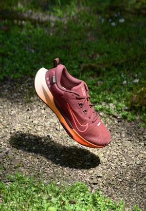 Rode en oranje hardloopschoen met Gore-Tex label zwevend boven een grindpad omgeven door groene planten en kleine witte bloemen.
