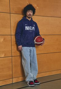 Violetinis gobtuvinis sportinis džemperis su baltu NBA logotipu, šviesiai mėlyni laisvi džinsai ir juodi/raudoni sportbačiai, laikantis raudoną krepšinį prieš medinę sieną.