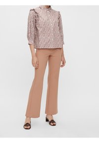 Blusa a fantasia floreale con dettagli arricciati sulle spalle, chiusura a bottoni e colletto. Abbinata a pantaloni a zampa d'elefante color pesca e sandali marroni.
