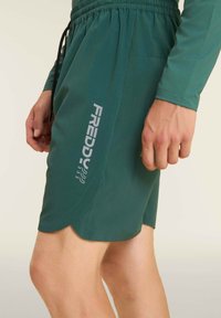 Pantalones cortos deportivos verdes con una cinturilla elástica, cordón de ajuste y un logo plateado en el lateral. Textura suave con un dobladillo ondulado.