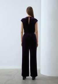 Lila sammetsoverall med korta ärmar, nyckelhål i ryggen och vida ben. Slät textur, figursydd midja och vanlig byxlängd.