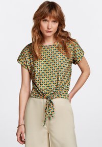 Lyhythihainen pusero vihreässä, oranssissa ja keltaisessa geometrisessä kuosissa. Ominaisuuksiin kuuluu solmittu yksityiskohta vyötäröllä ja pyöreä pääntie.