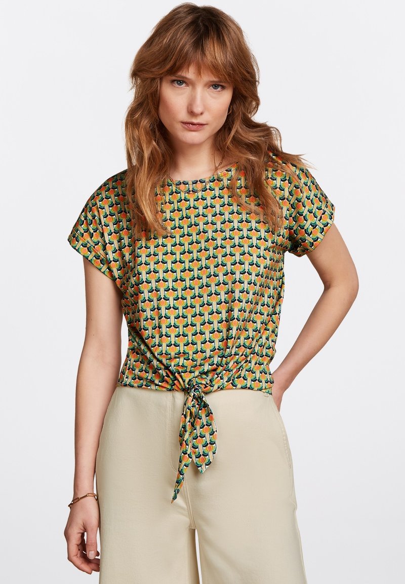 Lyhythihainen pusero vihreässä, oranssissa ja keltaisessa geometrisessä kuosissa. Ominaisuuksiin kuuluu solmittu yksityiskohta vyötäröllä ja pyöreä pääntie.