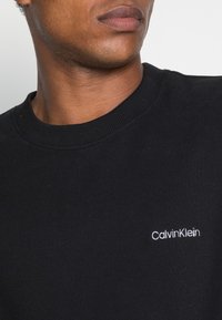 Felpa nera realizzata in morbido tessuto, con un colletto a coste e un piccolo logo bianco ricamato "Calvin Klein" sul petto.