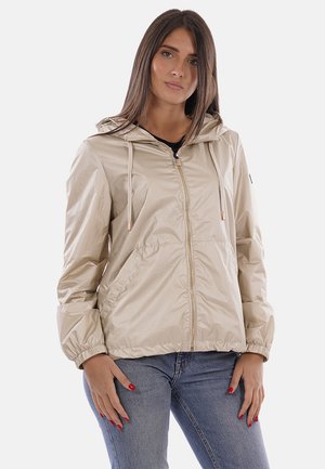 Donna che indossa una giacca beige con cappuccio e zip con cordini e jeans blu, in piedi con le mani rilassate lungo i fianchi.