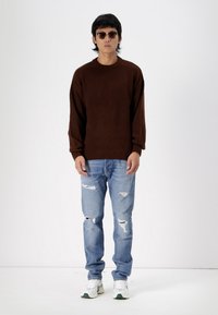 Brun strikket sweater, lyseblå slidte jeans og hvide sneakers med grønne accenter. Modellen står foran en ensfarvet baggrund, iført solbriller.