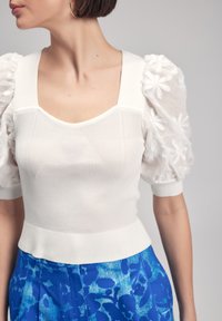 Top bianco lavorato a maglia con scollatura quadrata, corpetto testurizzato e maniche a palloncino trasparenti con motivo floreale; abbinato a pantaloni blu a fantasia.