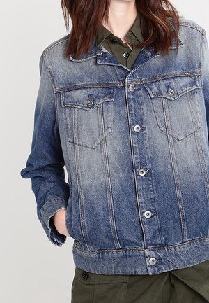 Kvinde iført en falmet blå denimjakke over en olivengrøn skjorte med knapper, med den ene hånd i jakke-lommen.