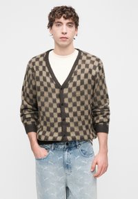 Bruin-beige geruit vest met V-hals, knoopsluiting en geribde manchetten, gecombineerd met lichtblauwe jeans met witte logo's.