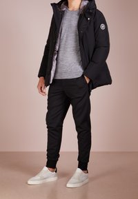 Osoba nosząca czarną kurtkę ocieplaną z futrzanym wykończeniem, szary sweter, czarne spodnie dresowe i białe sneakersy, stoi na beżowej podłodze.