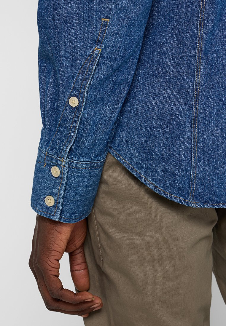 Chemise en denim à manches longues. Présente une couleur bleu clair, des poignets boutonnés et des ourlets cousus. Portée avec un pantalon beige.