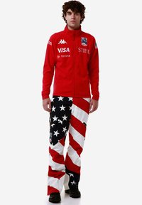 Uomo che indossa una giacca rossa della squadra USA con loghi di sponsor e pantaloni a fantasia con bandiera americana, in piedi su uno sfondo bianco.