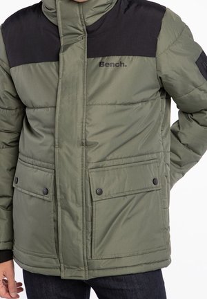 Zielona i czarna ocieplana kurtka typu bomber z przednimi kieszeniami na zatrzaski i zamkiem błyskawicznym, noszona przez osobę, której ręce są częściowo widoczne.