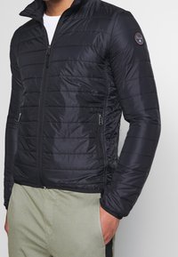 Schwarze Steppjacke mit horizontalem Qulitedesign, hohem Kragen, durchgehendem Reißverschluss und Seitentaschen. Hergestellt aus leichtem, reflektierendem Material.