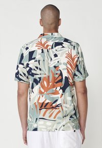 Camisa de manga corta con un estampado de hojas tropicales en tonos terrosos de verde, beige, naranja y marino, confeccionada en una tela ligera.