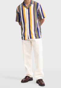 Chemise à manches courtes avec des rayures verticales multicolores en vert, jaune, violet et blanc. Associée à un pantalon de couleur crème.