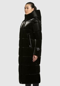Cappotto lungo imbottito nero con finitura lucida, colletto alto e tasche laterali con zip. Presenta un motivo a griglia cucito e un marchio ben visibile sulla manica.