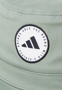 Zeleni klobuk iz blaga z teksturo, na katerem je okrogel črno-beli logotip s tremi črnimi črtami. Vidna šivanairka.