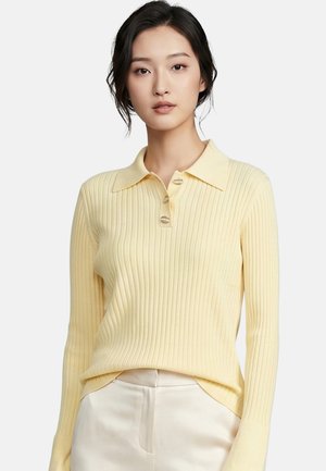 Donna che indossa un maglione polo giallo chiaro a coste con maniche lunghe, tre bottoni e pantaloni bianchi a vita alta su sfondo uniforme.