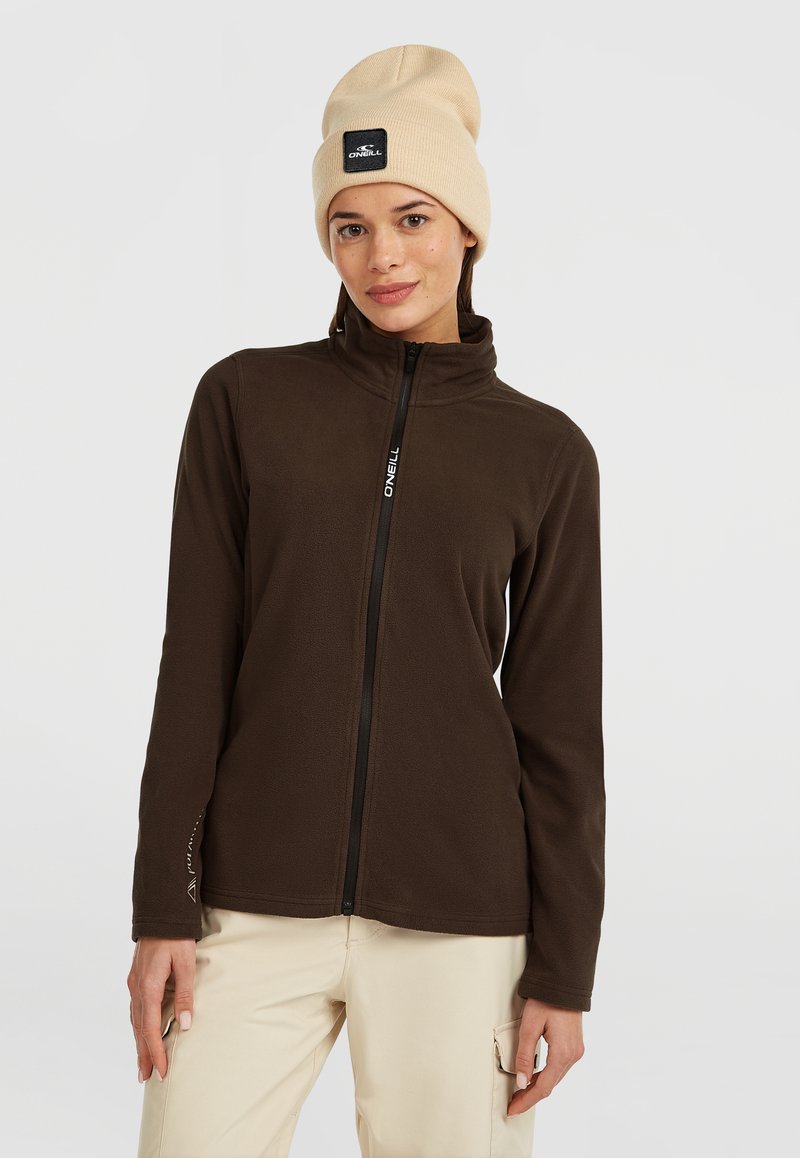 Giacca in pile marrone con colletto alto, chiusura con zip e dettagli con logo. Il modello indossa un berretto in maglia beige. Tessuto morbido, stile casual.